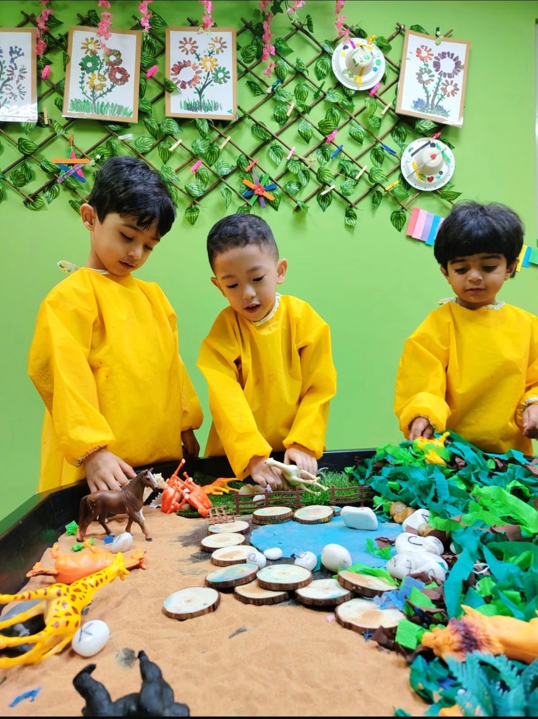 Three children in yellow smocks playing with toy animals in a sand and water tray